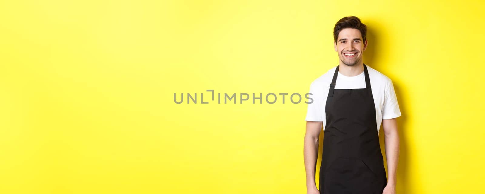 Young man waiter in black apron, smiling, working in store or coffee shop, standing against yellow background by Benzoix