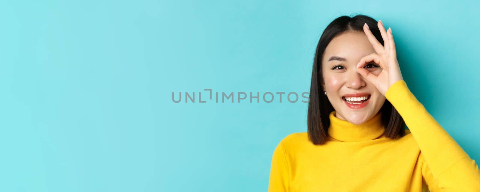 Beauty and makeup concept. Close up of carefree asian girl showing OK sign on eye and smiling, looking happy at camera, standing over blue background by Benzoix