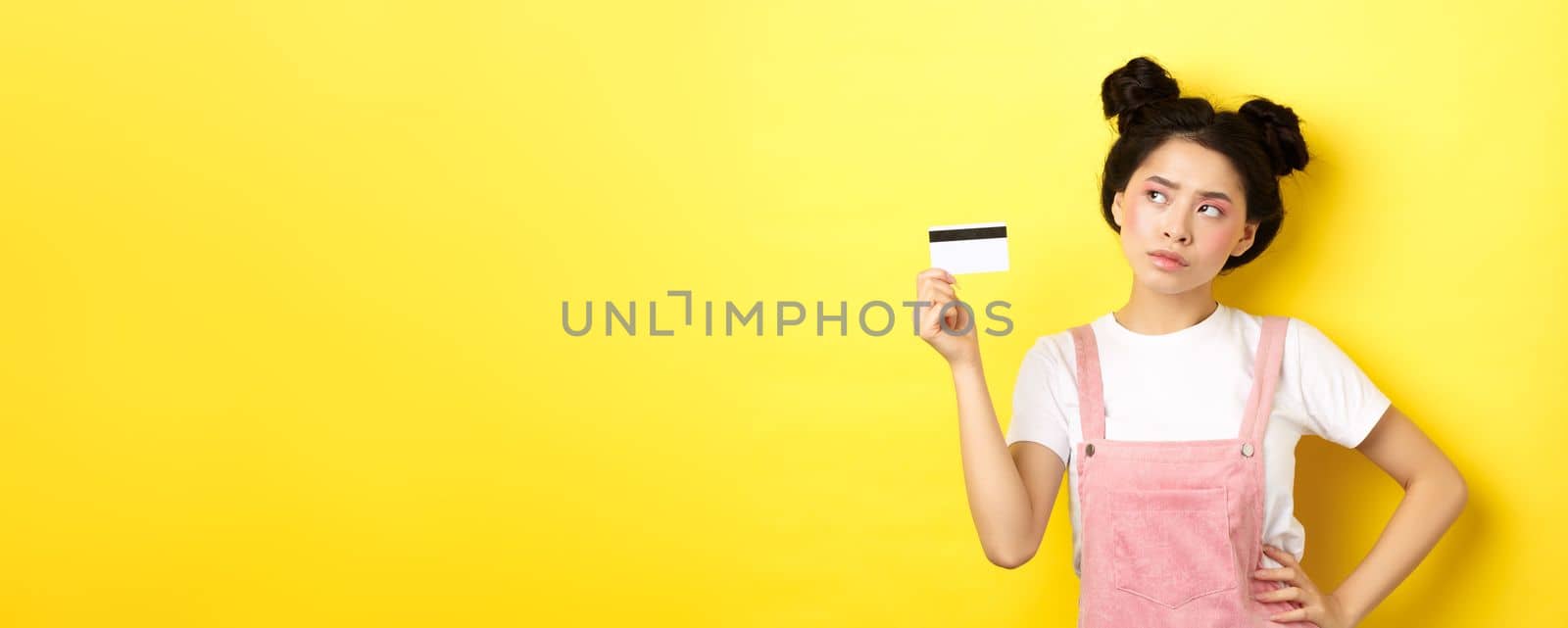 Shopping. Thoughtful asian woman with glamour makeup, holding plastic credit card and looking aside pensive, make decision, yellow background by Benzoix