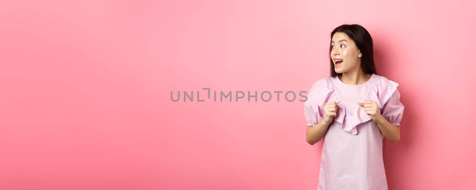 Excited asian girl look left with motivated face, smiling happy, standing in dress on pink background.