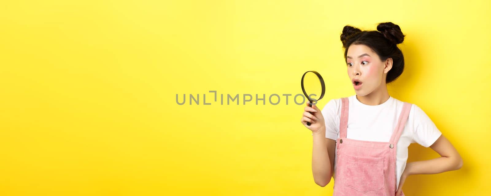 Excited asian woman with bright glamour makeup, look through magnifying glass amazed, staring aside, standing on yellow background by Benzoix