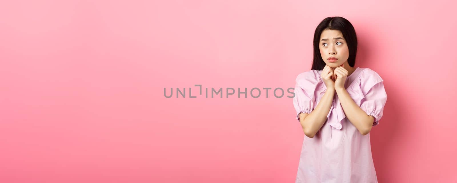 Worried timid asian girl looking aside, holding hands pressed to chest and look concerned, standing sad and gloomy on white background.