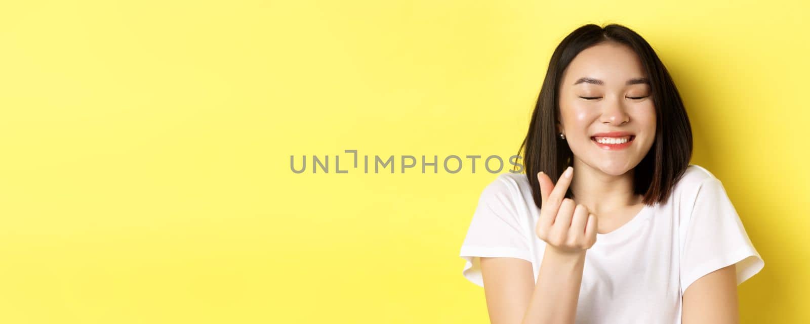 Valentines day and women concept. Close up of pretty asian girl in white t-shirt, smiling and showing finger heart, I love you gesture, standing over yellow background.