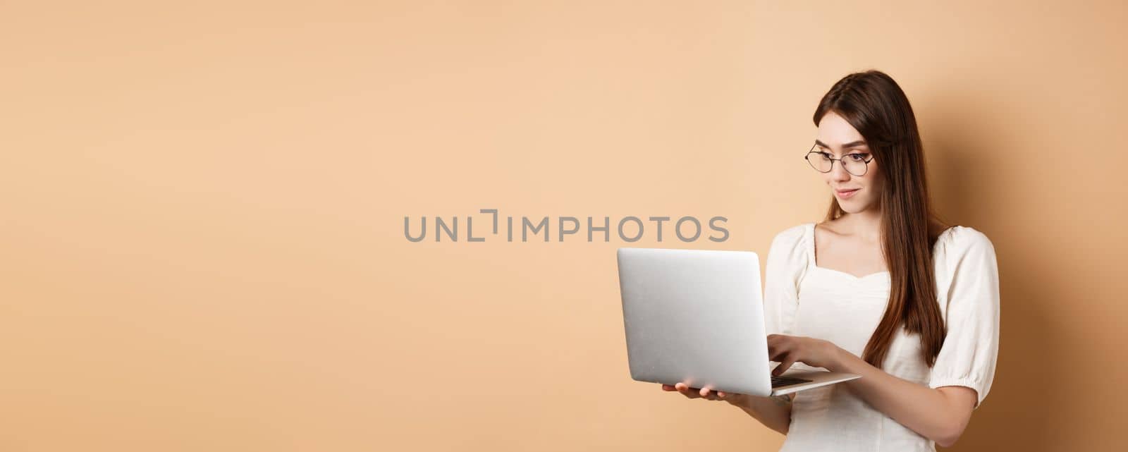 Young woman in glasses working on laptop, reading computer screen and smiling, standing on beige background by Benzoix