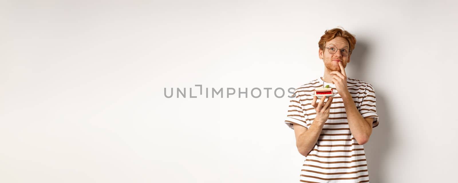 Holidays and celebration concept. Happy young man with red hair and glasses having birthday, holding cake with candle and thinking of b-day wish, standing over white background by Benzoix