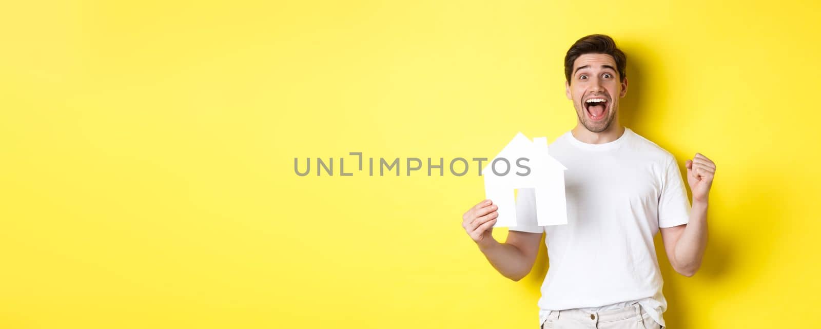 Real estate concept. Excited man holding paper house model and celebrating, standing happy over yellow background.