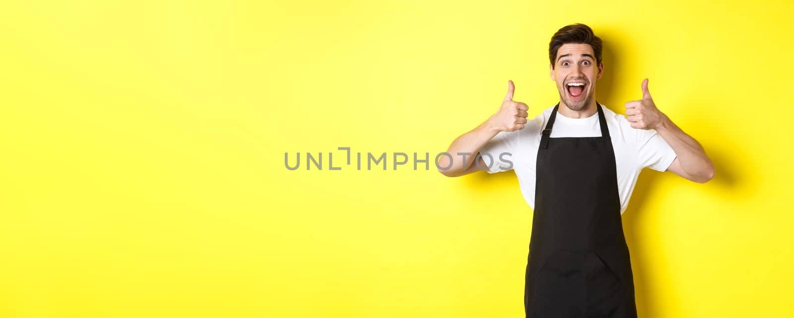 Cheerful man barista in black apron showing thumbs-up, recommending cafe or restaurant, standing against yellow background by Benzoix