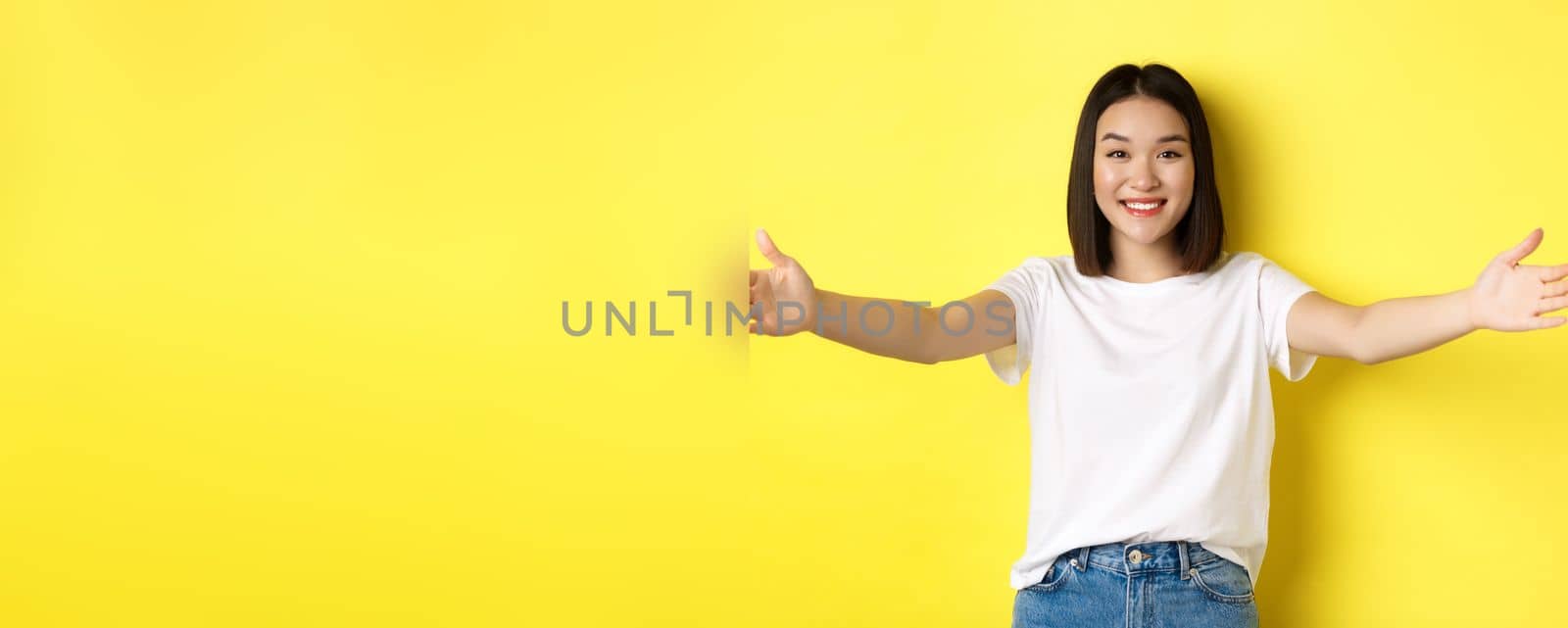 Beauty and fashion concept. Friendly asian woman spread out hands and smiling, waiting for hugs, inviting you, welcome someone and looking happy, standing over yellow background.