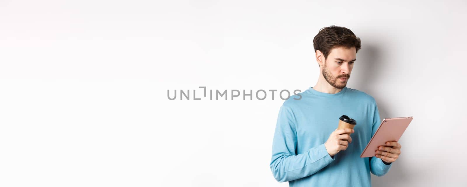 Man reading news online in digital tablet, looking serious at screen while drinking coffee from paper cup, standing over white background.