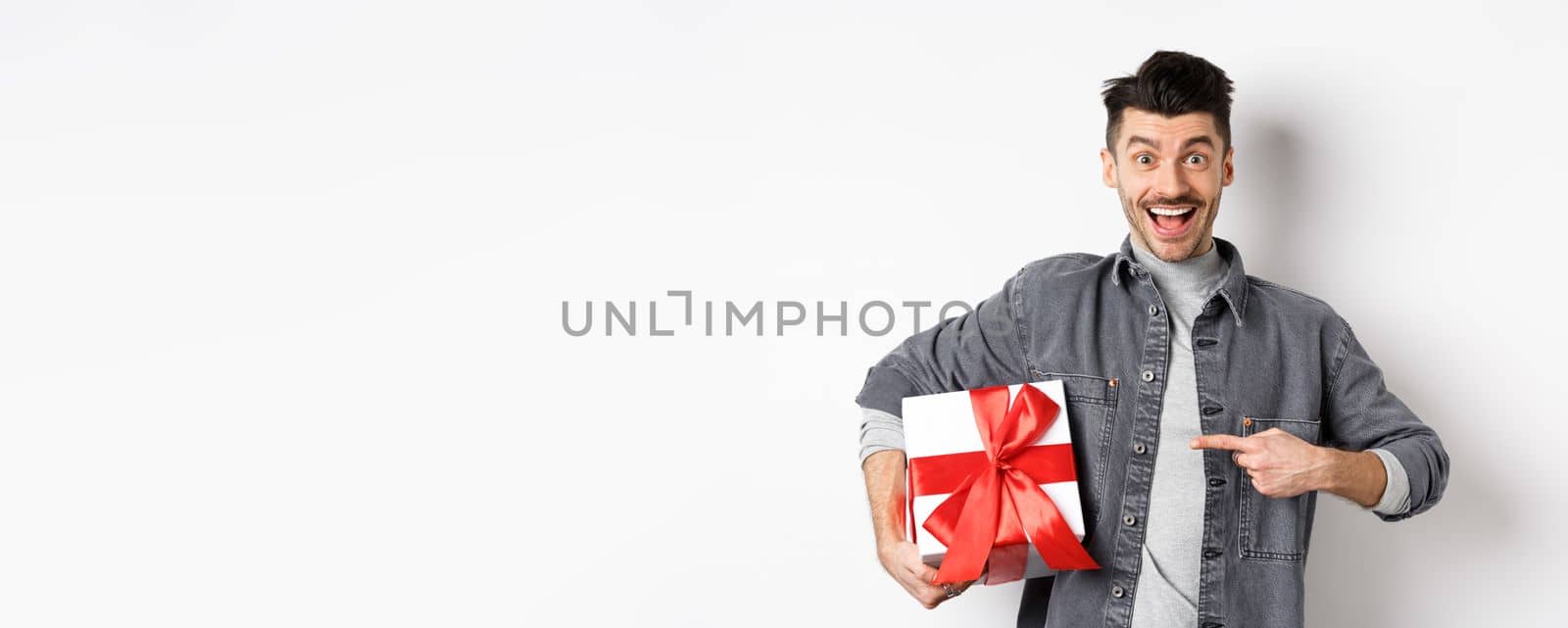 Happy Valentines day. Excited caucasian guy pointing at gift box from lover, smiling and looking amazed, buying presents on romantic date, white background by Benzoix