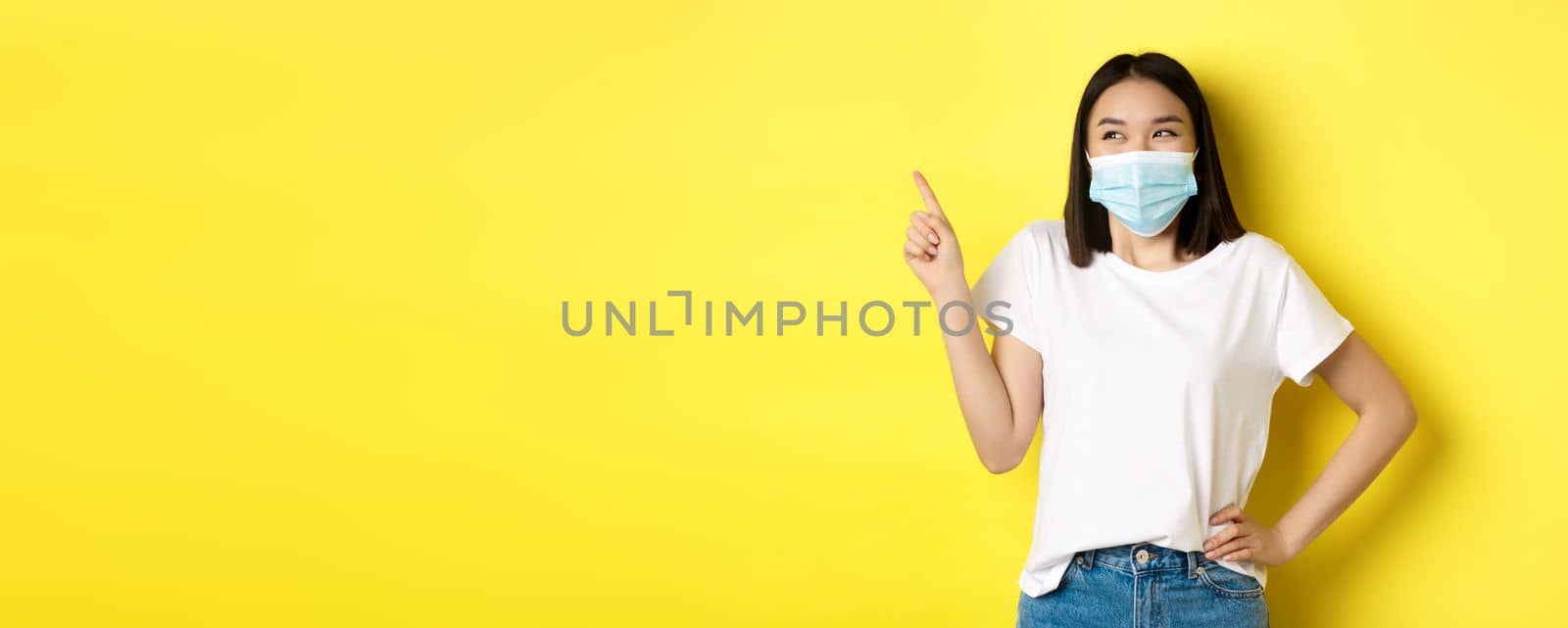 Covid, health care and pandemic concept. Asian woman in medical mask and white t-shirt pointing finger at upper left corner logo, showing promotion, yellow background.
