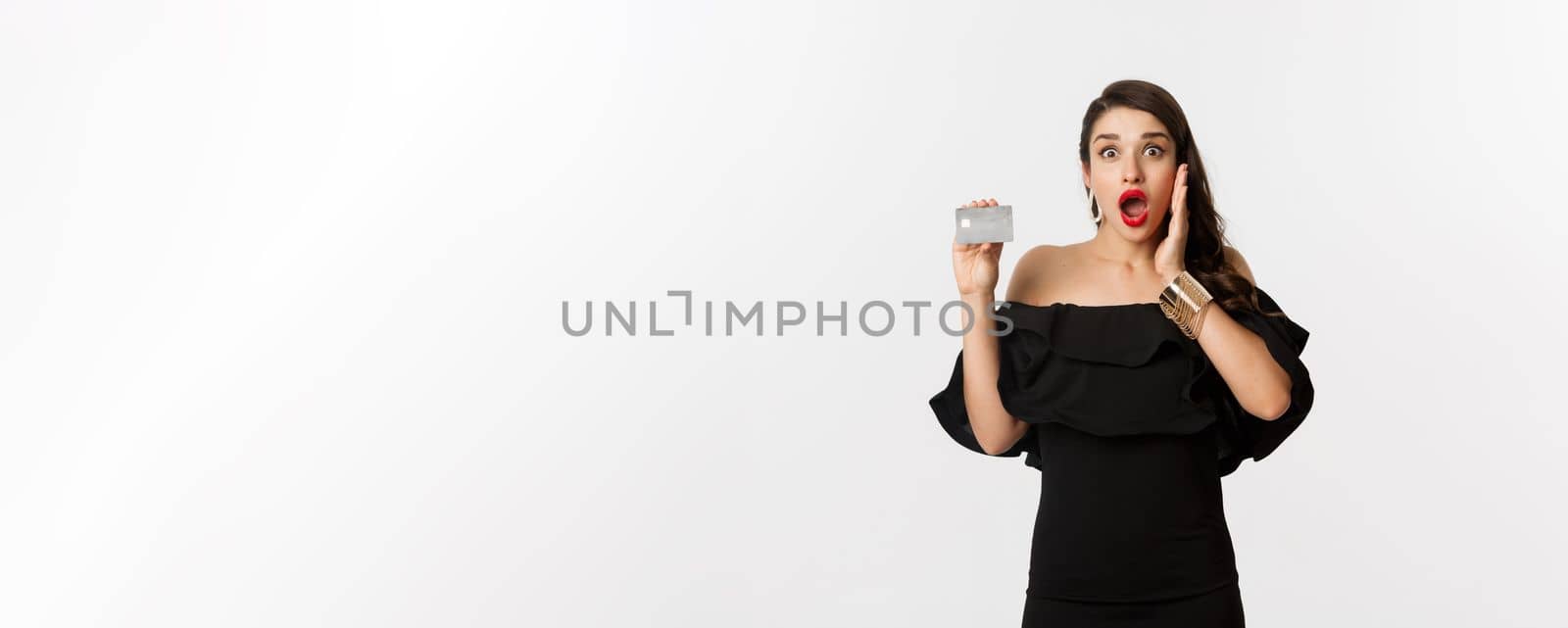 Fashion and shopping concept. Excited elegant woman with red lips, wearing black dress, showing credit card and looking amazed, standing over white background by Benzoix