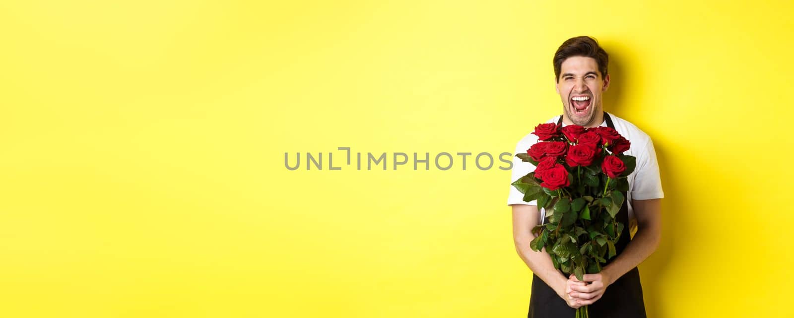 Funny young salesman in black apron holding bouquet of roses, florist laughing and standing over yellow background.