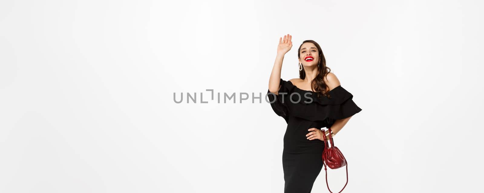 Beauty and fashion concept. Full length of attractive young woman in black dress and makeup, saying hello and smiling, waving hand to greet, white background by Benzoix
