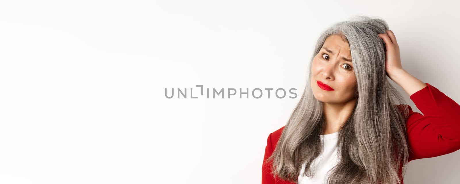 Close up portrait of confused asian female manager scratching head and looking questioned at camera, standing over white background by Benzoix
