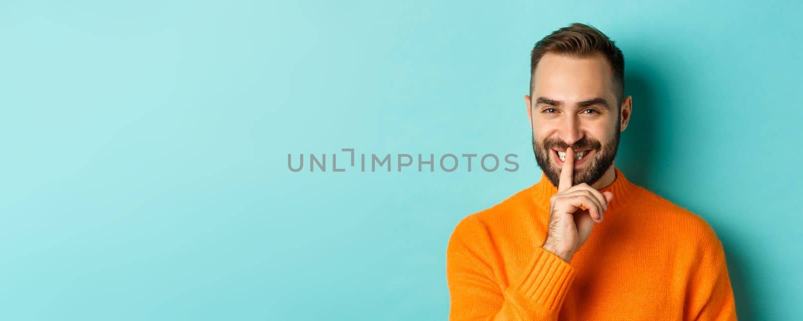 Close-up of handsome smiling man hushing, telling secret, standing over light blue background.