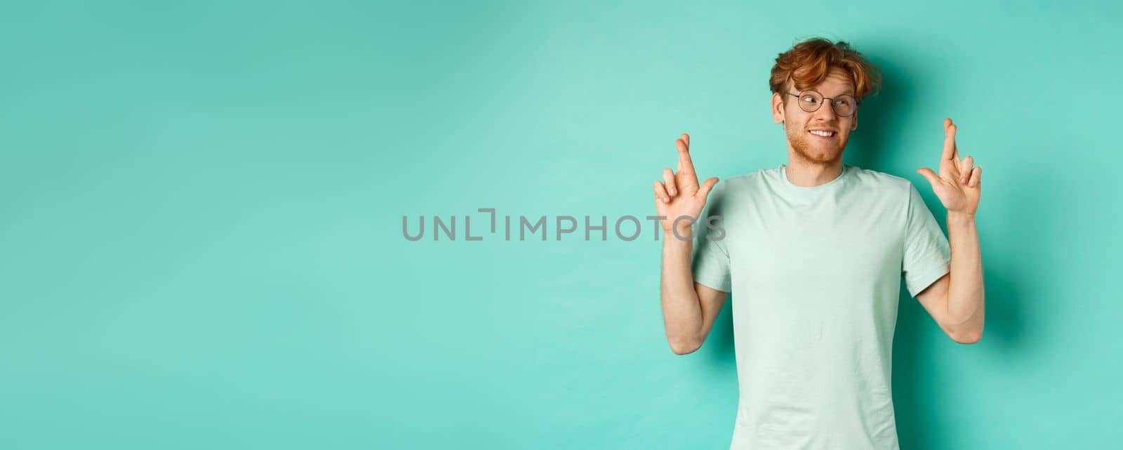 Hopeful redhead man in glasses making a wish, cross fingers and looking right at copy space, dreaming about something or making wish, standing over turquoise background.