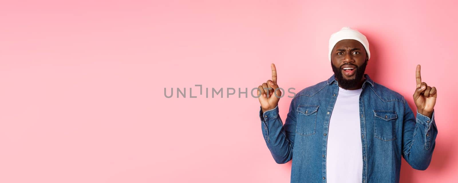 Disappointed and upset Black man frowning, pointing fingers up and staring at camera bothered, complaining at something, standing over pink background by Benzoix