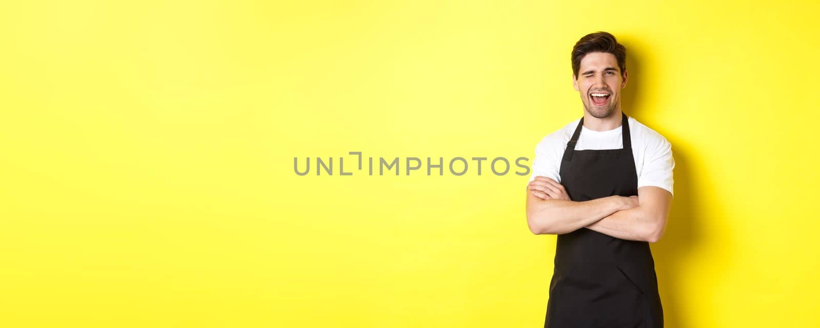 Handsome barista in black apron winking at you, wearing black apron uniform, standing over yellow background.