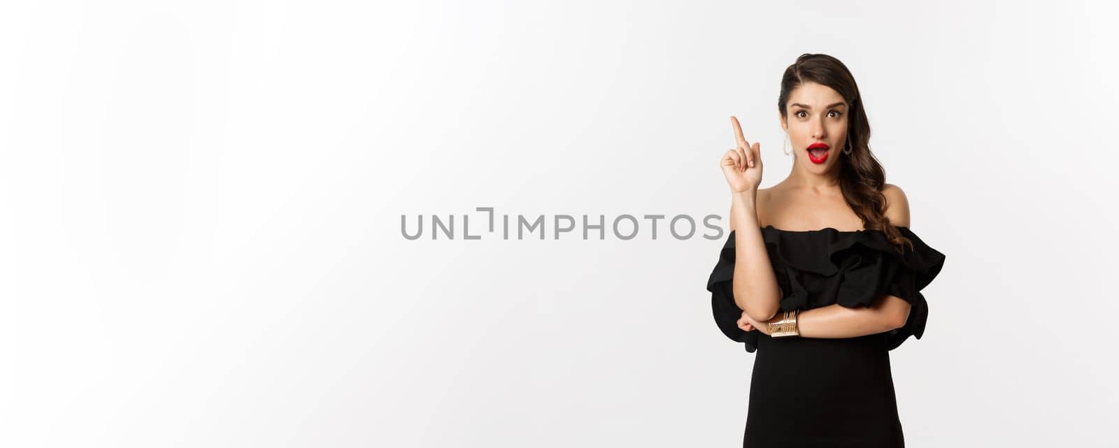 Fashion and beauty. Attractive caucasian woman in black dress having an idea, raising finger and saying suggestion, white background.