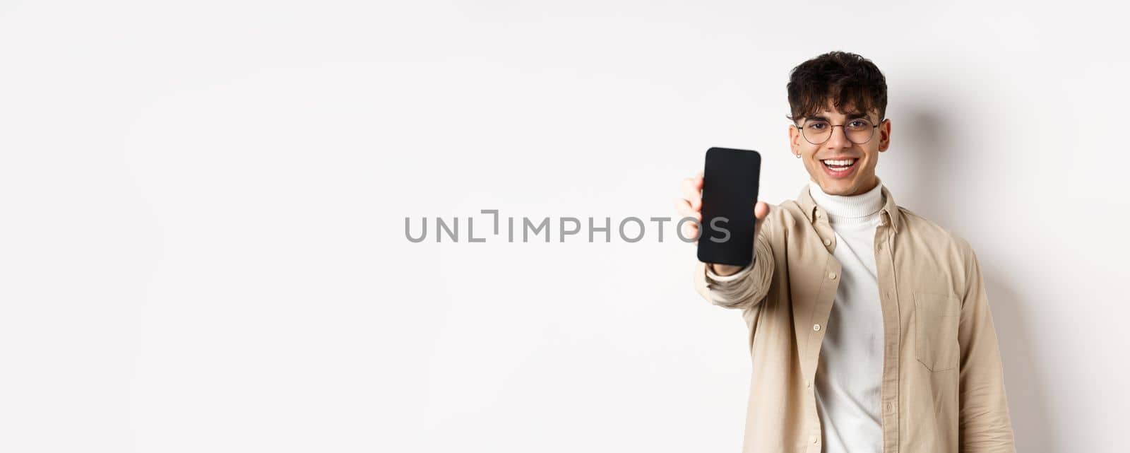 Handsome young man showing empty smartphone screen, standing on white background. Copy space