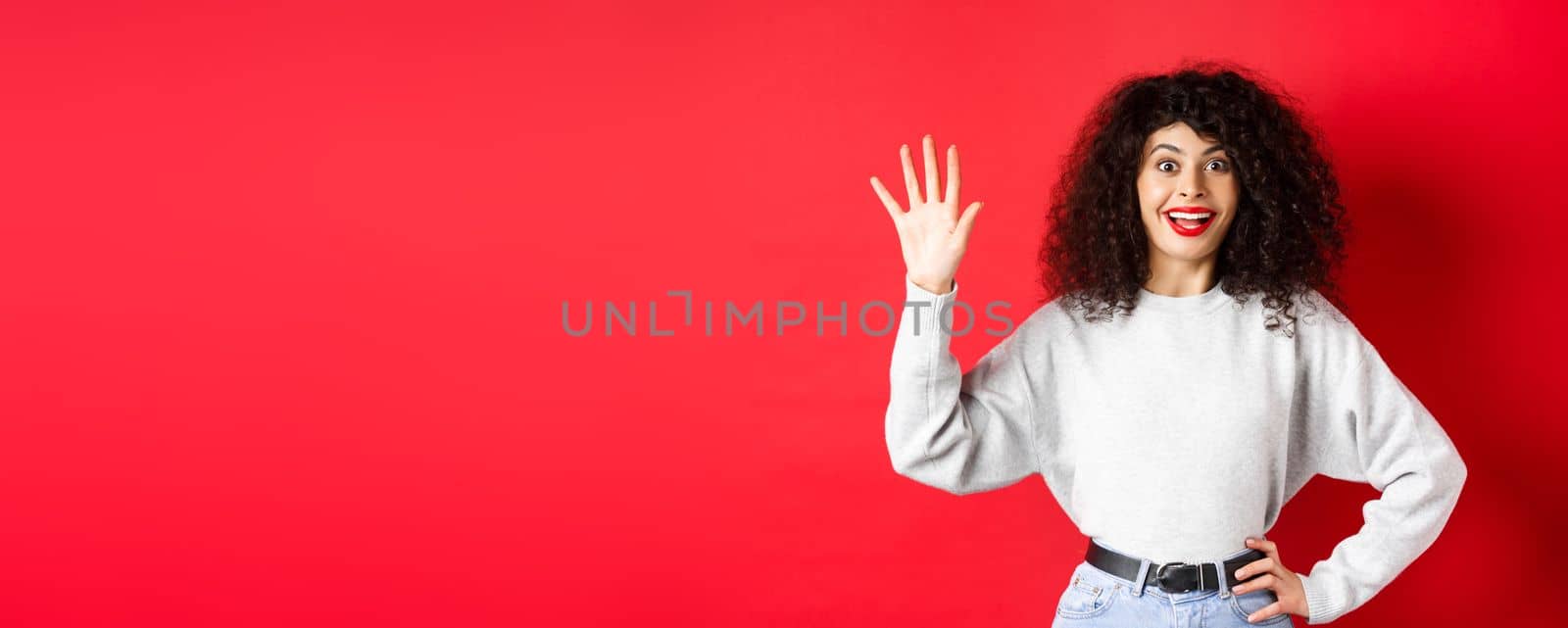 Excited woman with curly hair showing number five with fingers, making order, standing against red background.
