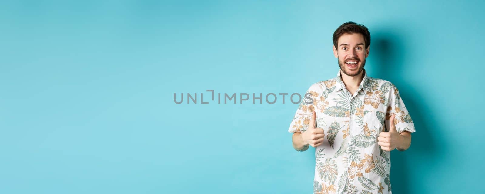 Cheerful guy showing thumbs up on vacation, praising travel agency, standing in summer shirt on blue background.