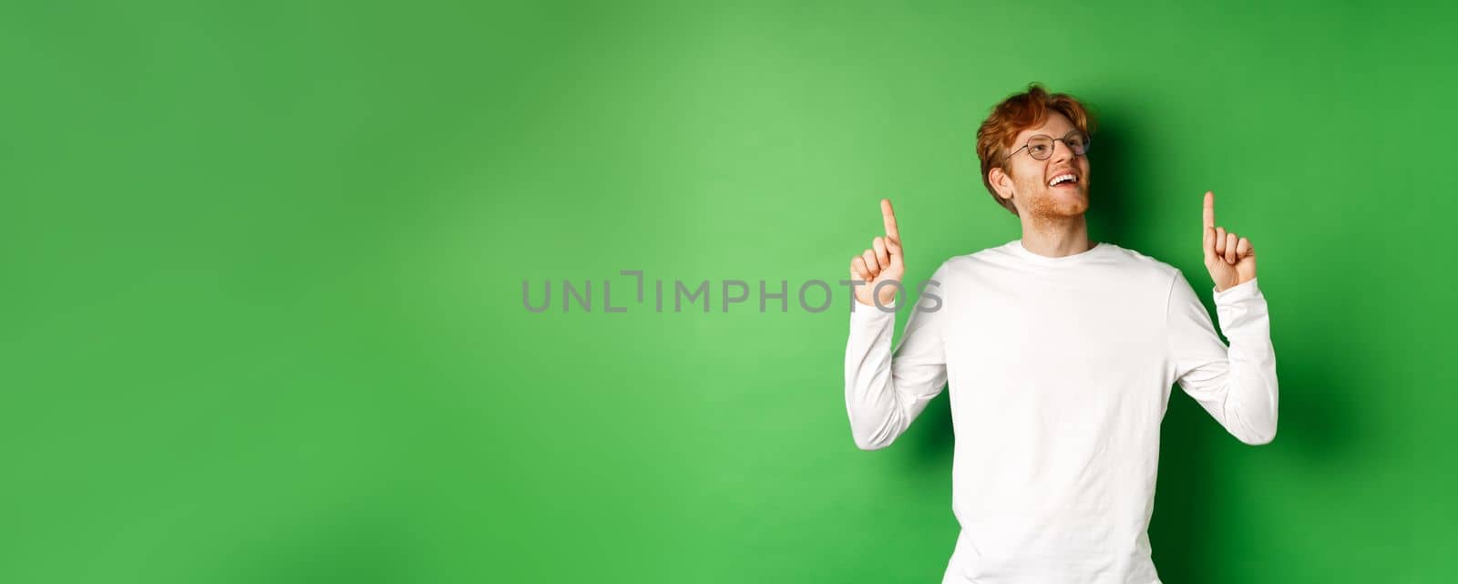 Cheerful handsome man in glasses checking out sale deal, pointing and looking up with happy and dreamy smile, standing over green background.