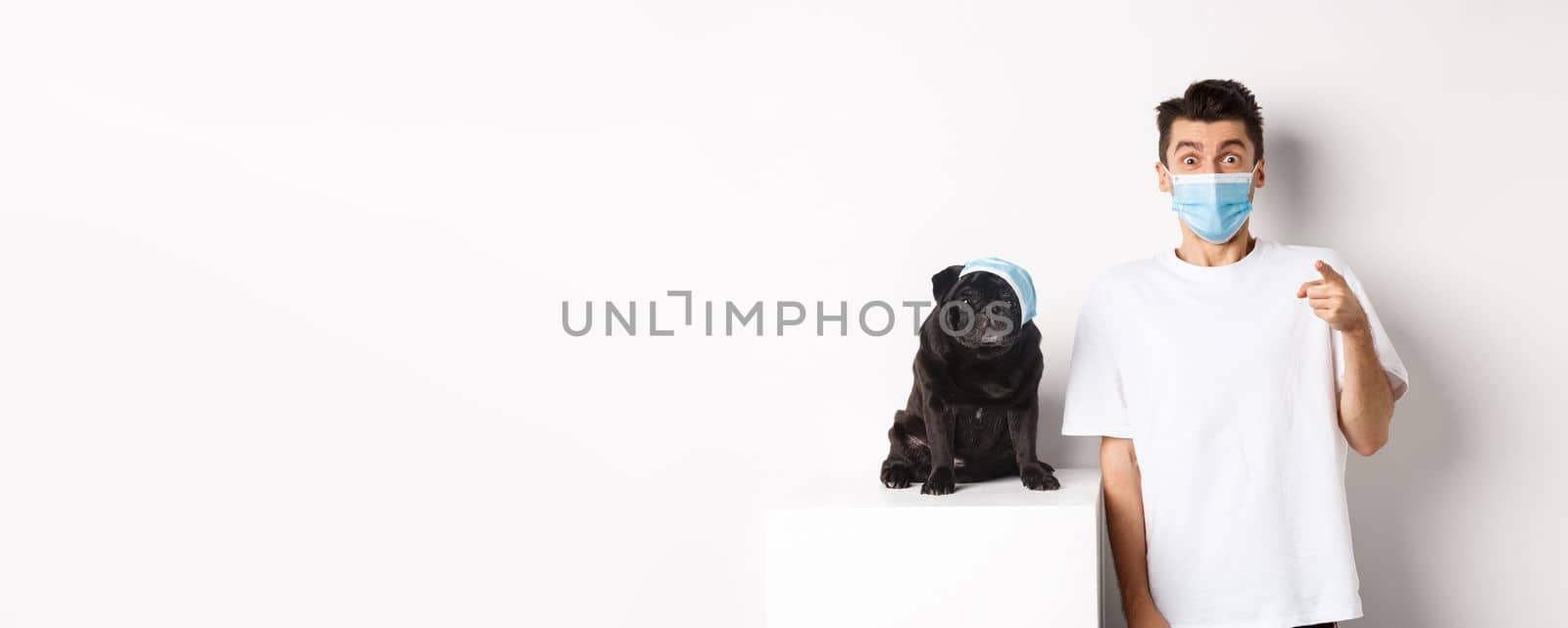 Covid-19, animals and quarantine concept. Happy dog owner and cute pug wearing medical masks, man pointing finger at camera amazed, white background by Benzoix