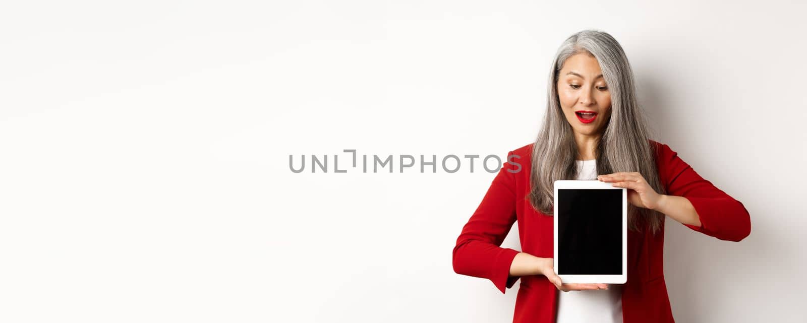 Business. Mature asian office lady showing digital tablet screen and looking down amazed, standing over white background by Benzoix