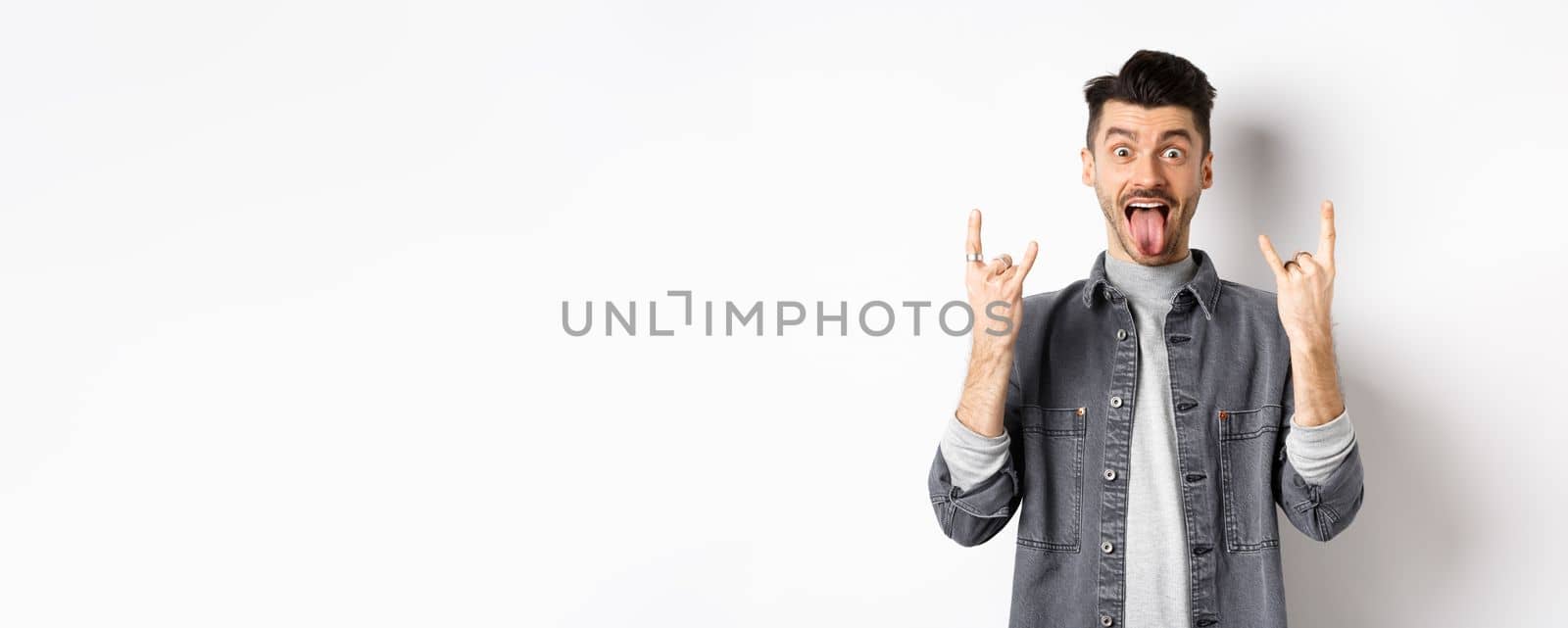 Excited funny guy showing tongue and heavy metal horns sign, enjoying party or event, having fun, standing on white background.