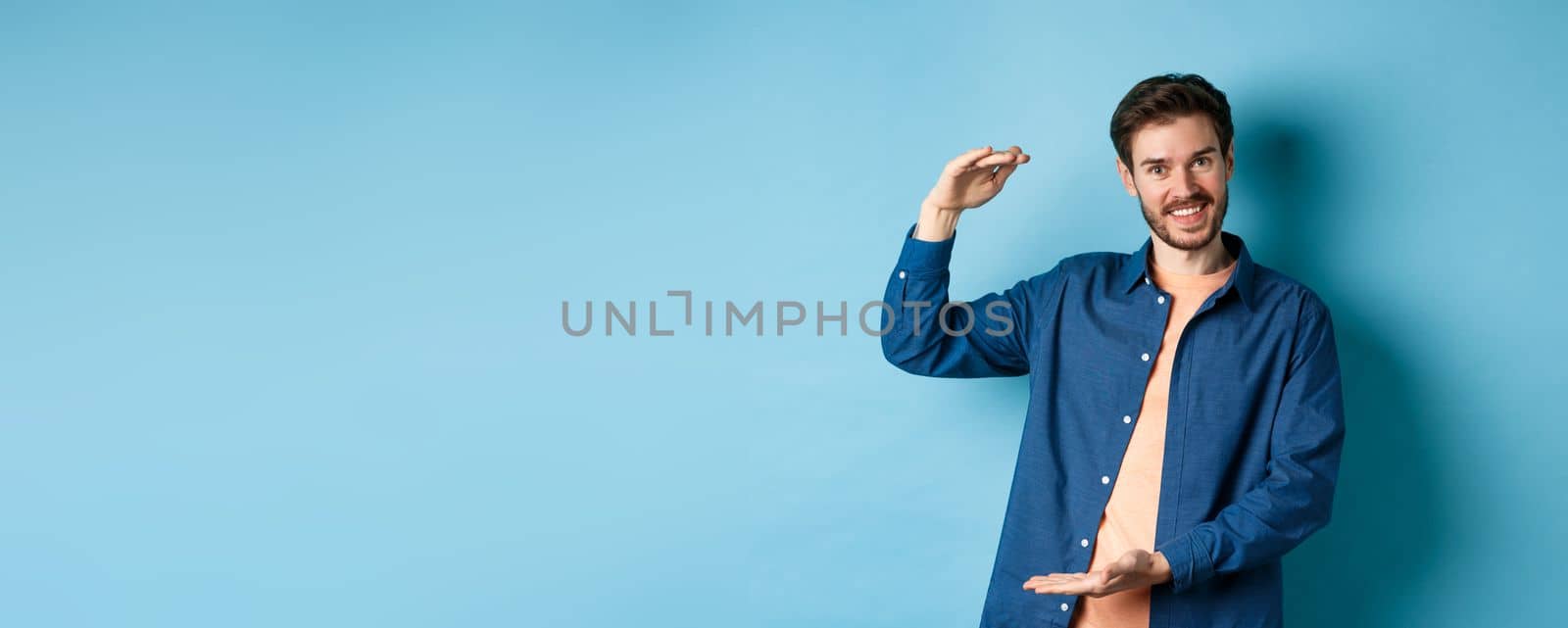 Handsome caucasian guy showing something big, shaping large size object with hands and smiling, standing on blue background by Benzoix