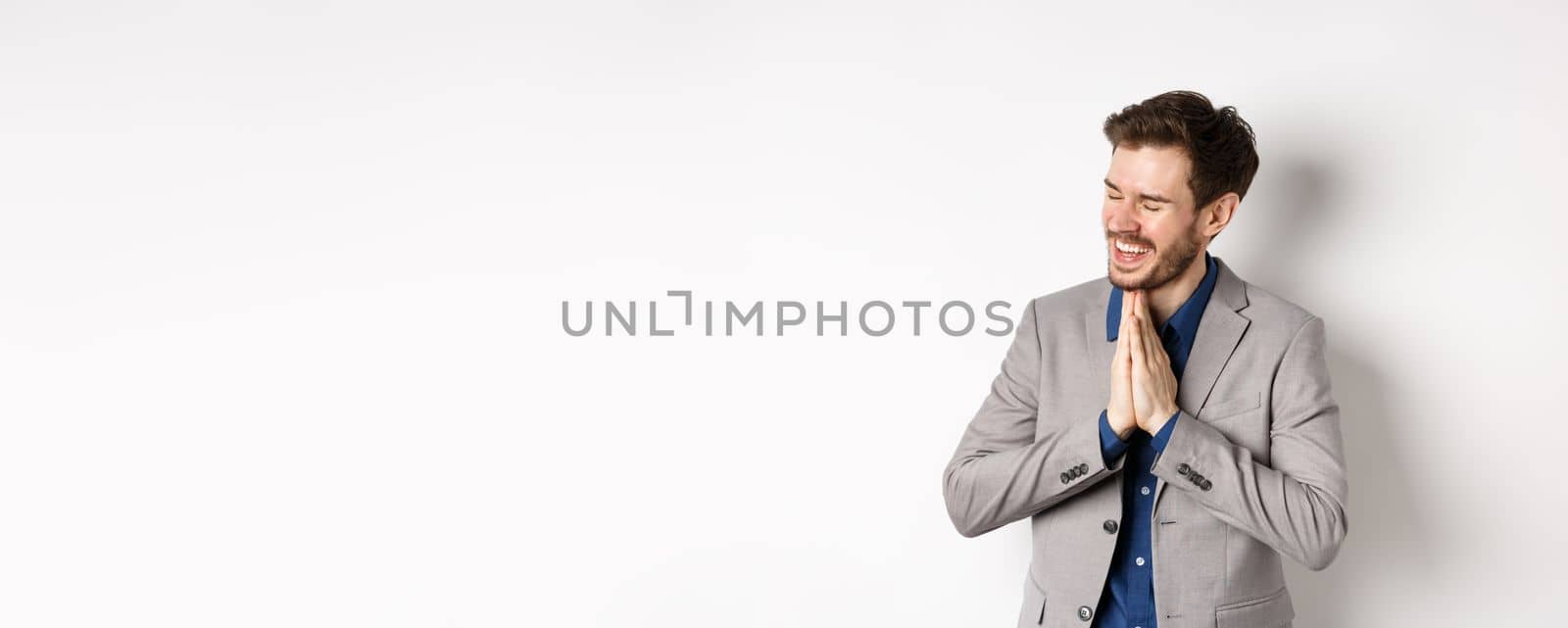 Happy and thankful businessman rejoicing, holding hands in begging or namaste sign, thanking for help, smiling relieved, standing in suit on white background.