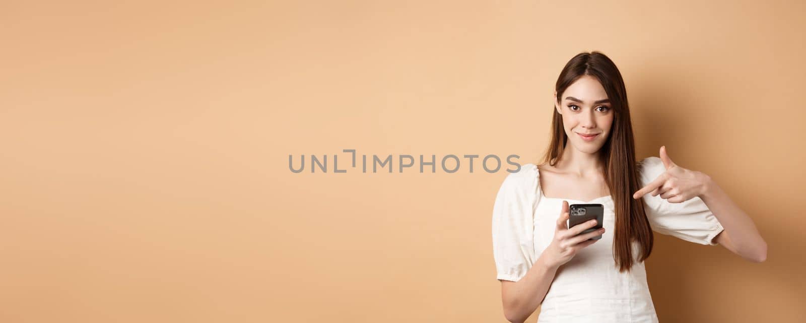 Attractive woman smiling and pointing at smartphone, showing online offer on phone, standing on beige background by Benzoix