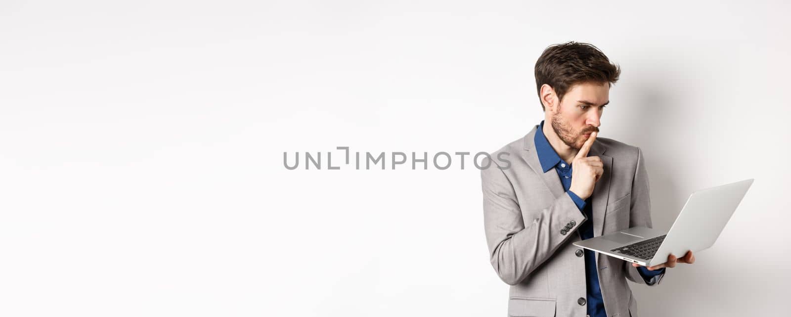Pensive businessman working on laptop and thinking, looking thoughtful at computer screen, standing in suit on white background.