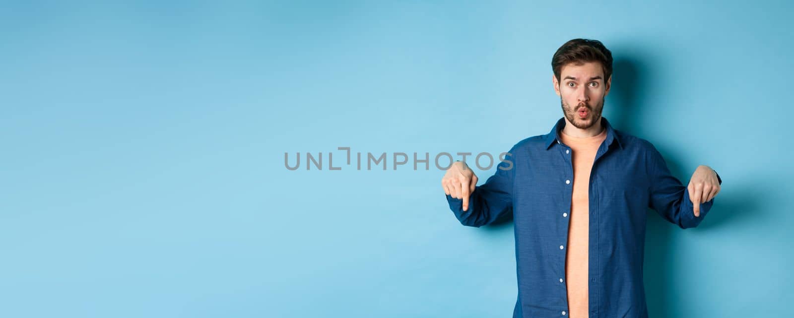 Impressed guy look curious, say wow and pointing fingers down, showing advertisement, standing on blue background.
