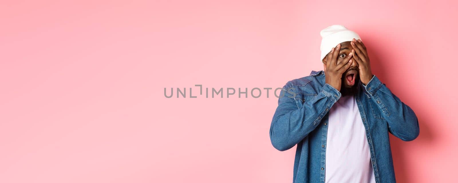 Curious Black man cover eyes but peeking through fingers, staring at camera amazed, standing in hipster beanie against pink background by Benzoix