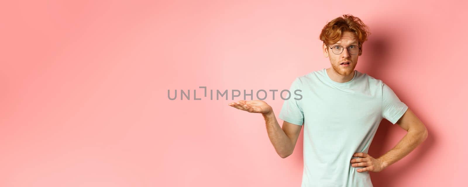 So what. Confused redhead man raising hand and shrugging, looking puzzled, dont understand something, standing over pink background.