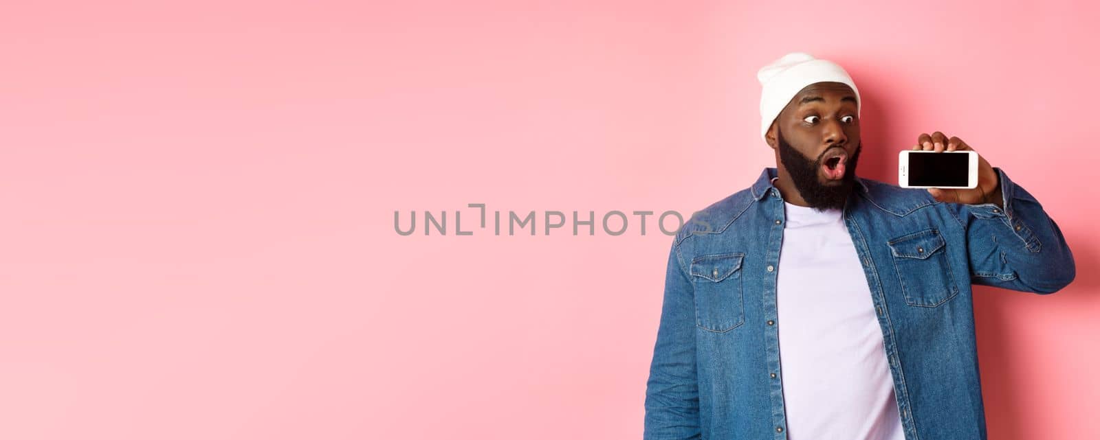 Online shopping and technology concept. Impressed african-american man in beanie showing phone screen, staring at mobile amazed, standing over pink background by Benzoix