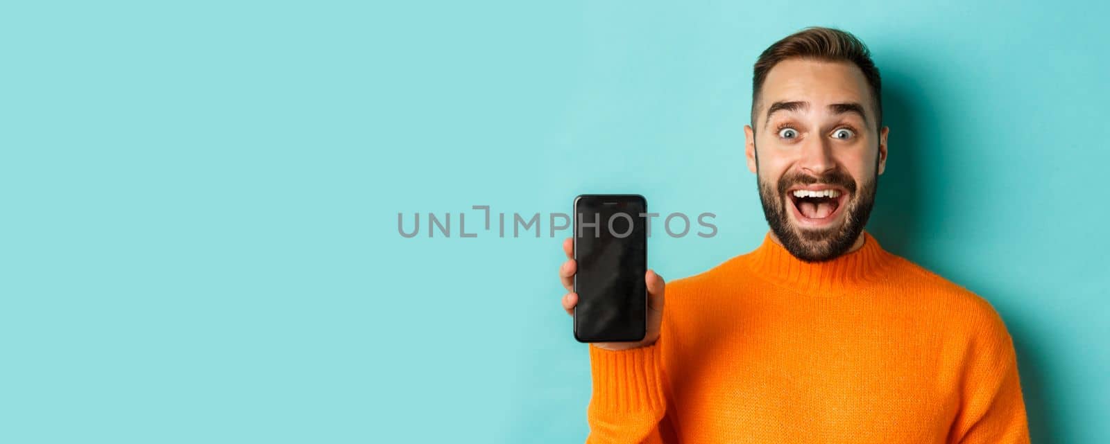Close-up of handsome bearded guy in orange sweater, showing smartphone screen and smiling, showing promo online, turquoise background.