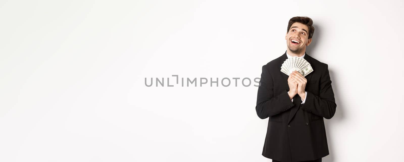 Image of handsome dreamy man in black suit, holding money and looking at upper left corner, thinking about shopping, standing over white background by Benzoix