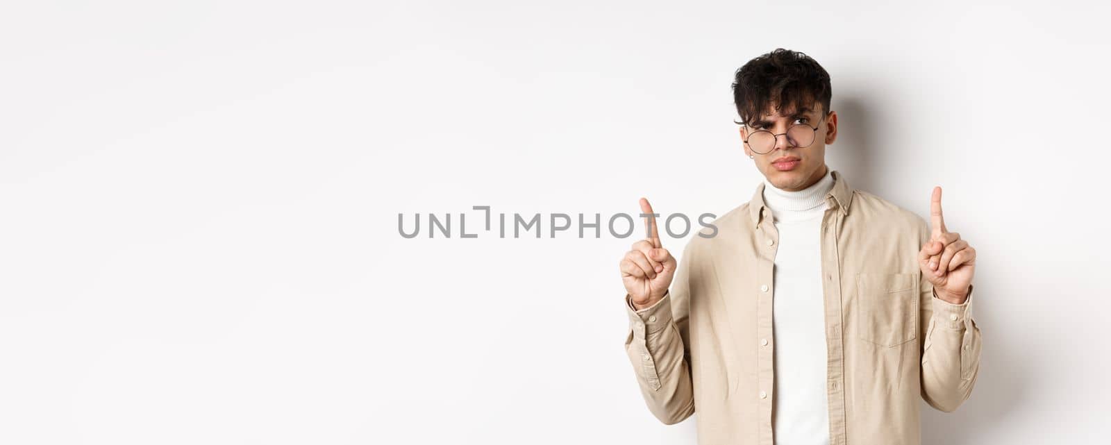 Portrait of young male model in glasses looking suspicious and frowning, pointing fingers up with disbelief, standing on white background.