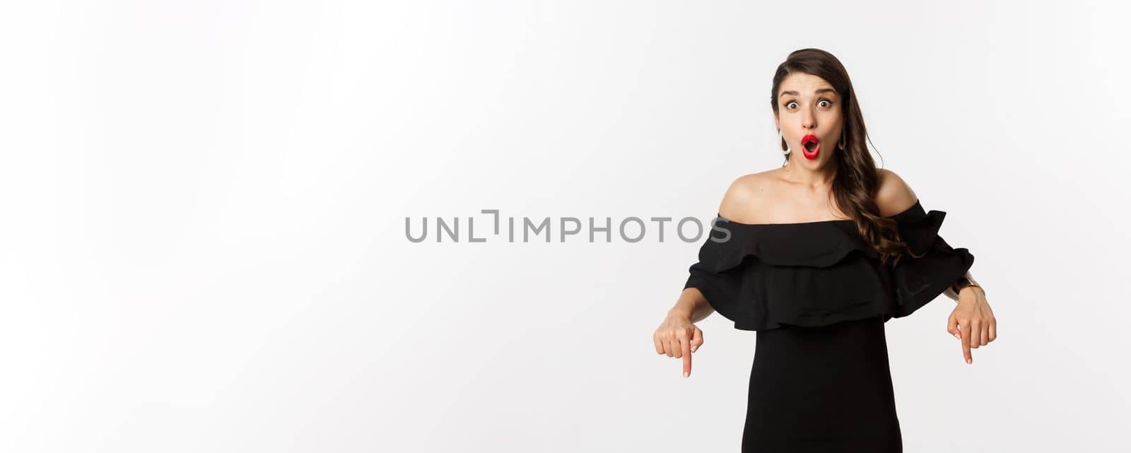 Fashion and beauty. Elegant woman in black dress pointing fingers down, showing promo and smiling, standing over white background by Benzoix