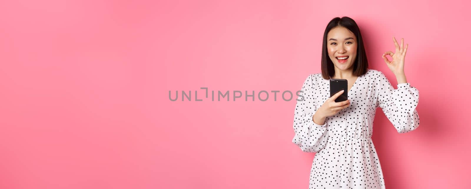 Online shopping and beauty concept. Satisfied asian female customer showing okay, making purchase in internet on smartphone, standing over pink background.