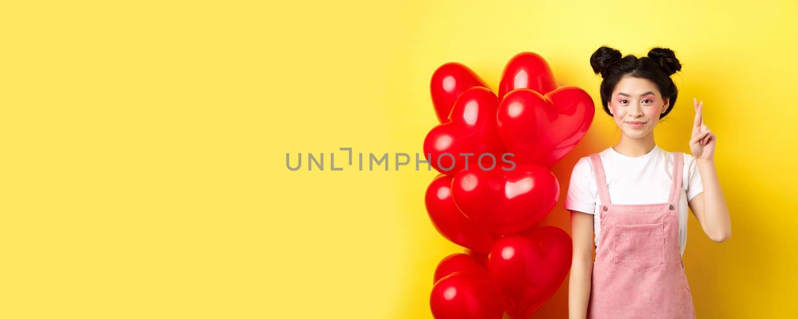 Valentines day concept. Stylish teenage asian girl with romantic make-up look, holding fingers crossed for good luck, making wish and smiling, standing on yellow background.