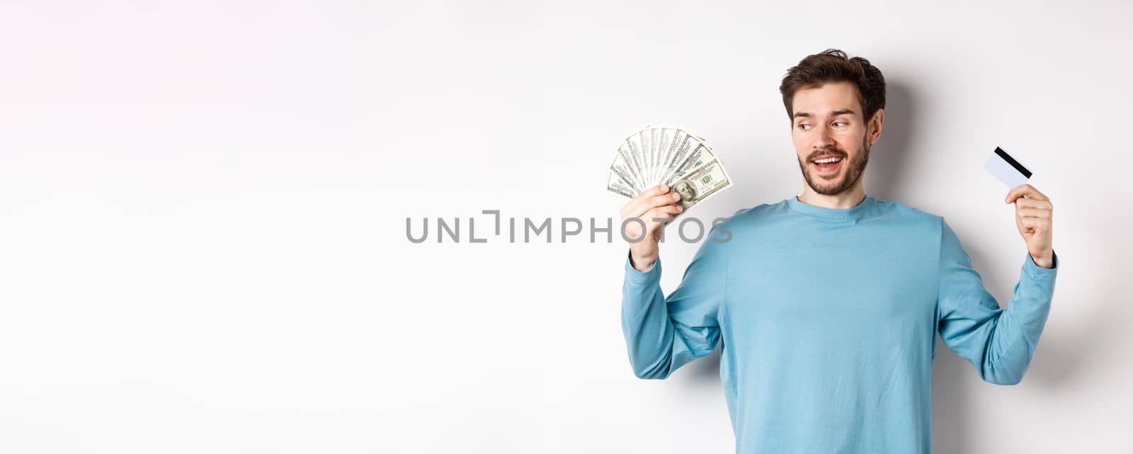 Excited bearded man choosing between money and plastic credit card, payment with cash or contactless, standing over white background by Benzoix