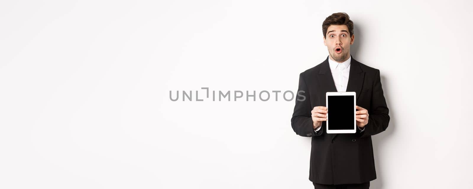 Image of surprised handsome man in black suit, showing digital tablet screen and looking amazed, standing against white background.