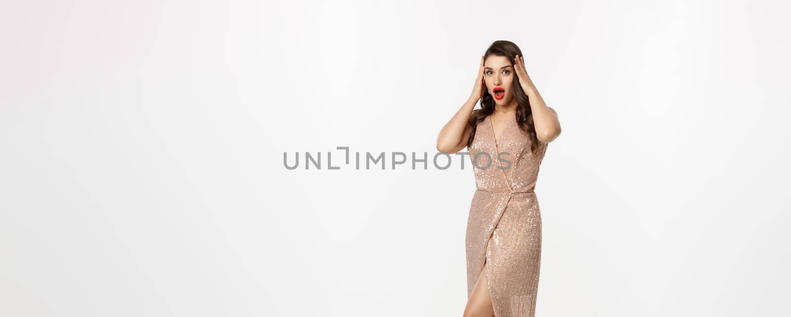 Christmas party and celebration concept. Full length of beautiful brunette woman looking surprised, wearing elegant dressed and staring with disbelief at camera, white background.