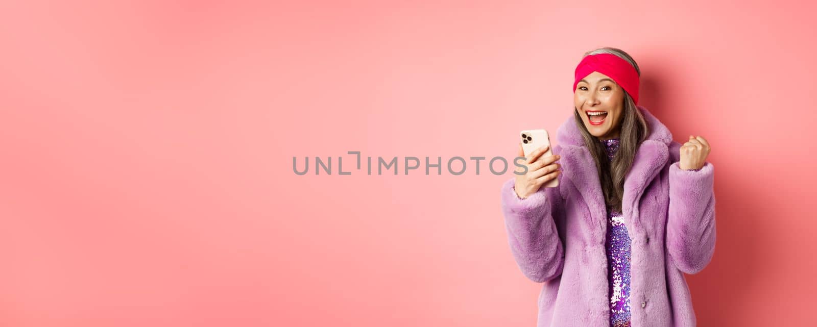 Online shopping and fashion concept. Happy asian senior woman winning prize in internet, holding mobile phone and making fist pump, scream of joy, standing over pink background by Benzoix