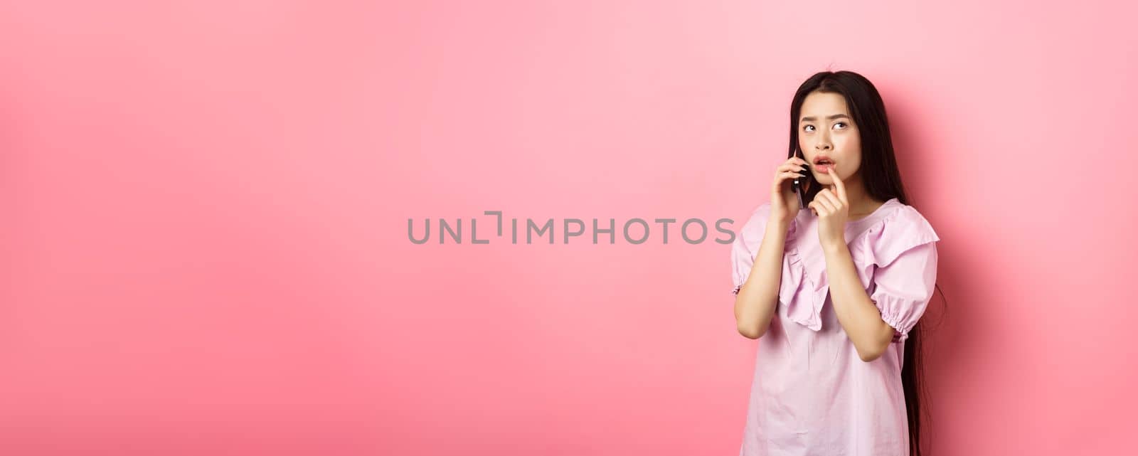 Thoughtful asian woman talking on mobile phone and thinking, touching lip and look up pensive, standing against pink background.